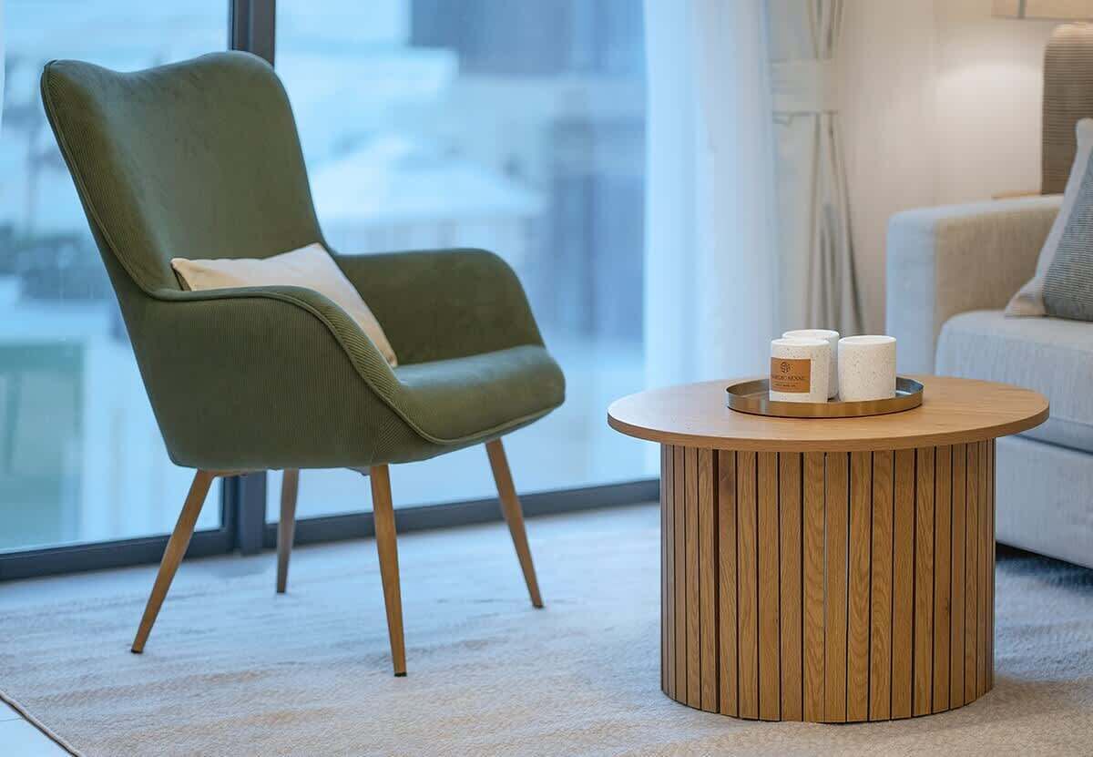A modern room with a green upholstered chair, a round wooden coffee table, and a view through a large window.