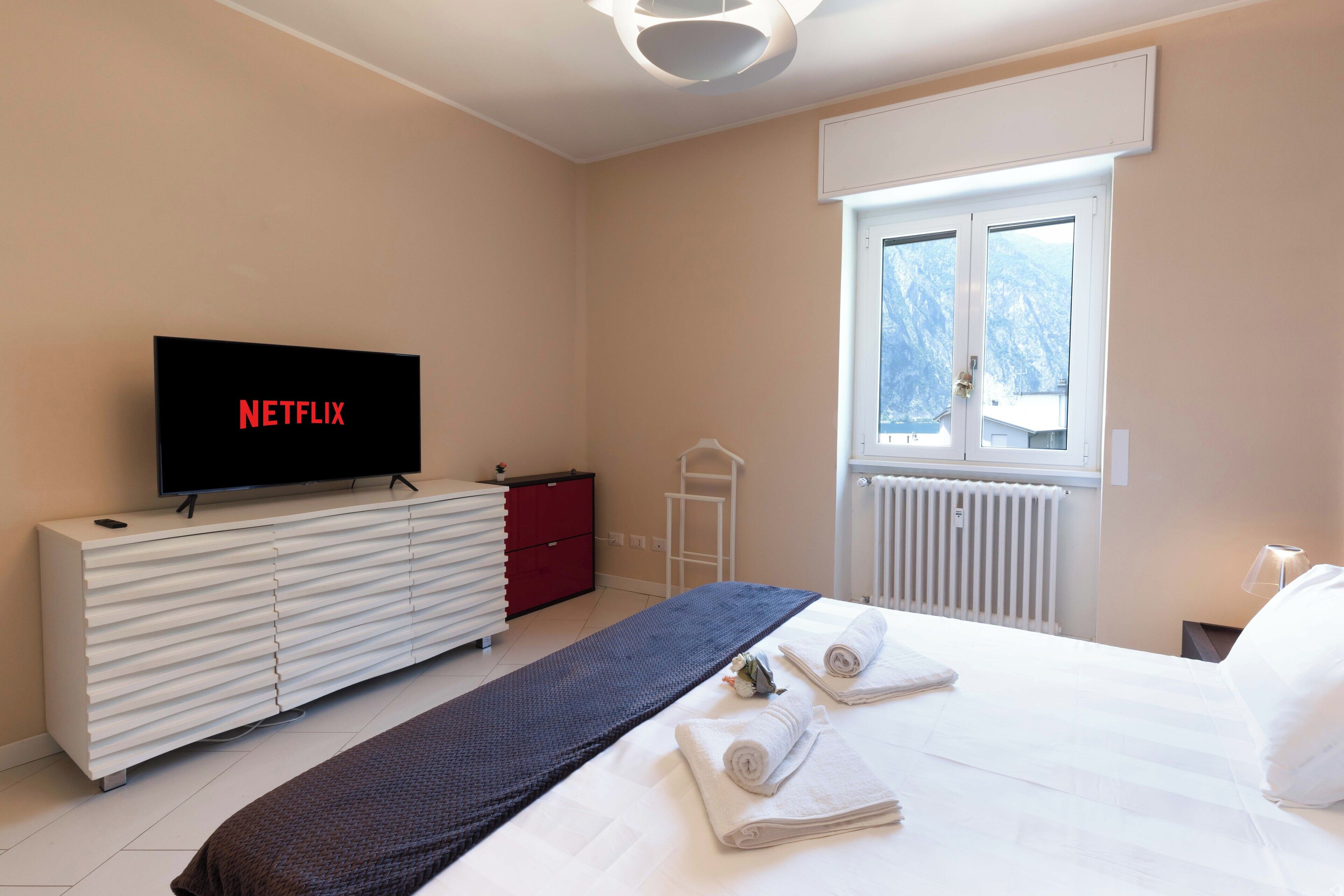 A hotel room with a bed, a TV on a white dresser, a window with a view, and a radiator.