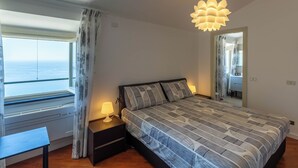 Un dormitorio con cama, lámparas de noche, ventana con cortinas y vista al mar.