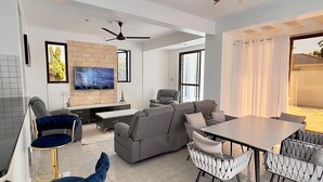 Een moderne woonkamer met een bank, een eettafel en barstoelen. Er is een televisie aan de muur en een plafondventilator.