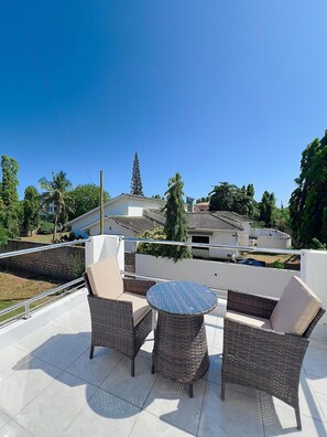A rooftop patio with wicker furniture, a round table, and a view of surrounding buildings and trees. - Elkhart Fereinhaus Villa (Mombasa)
