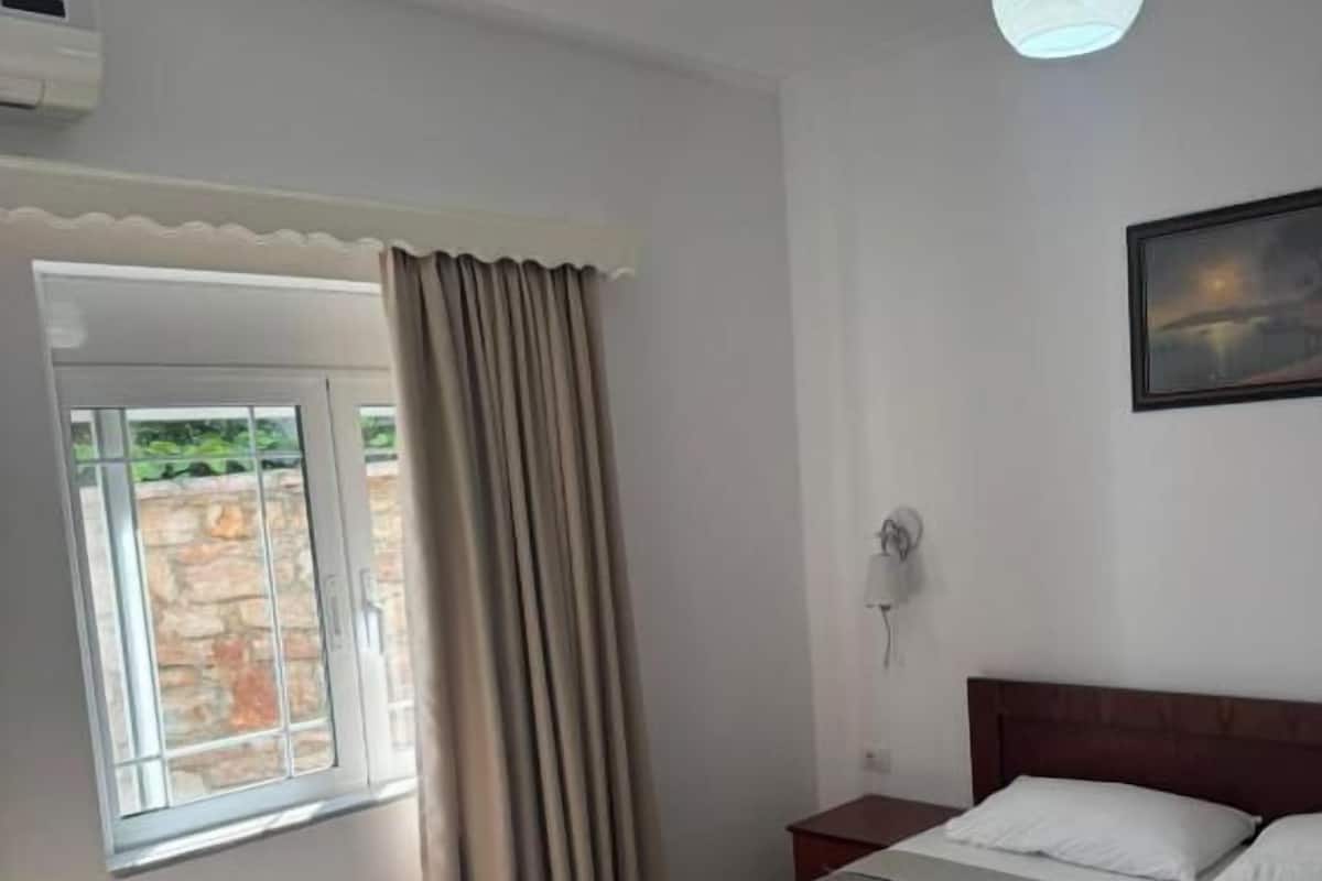 A hotel room with a bed, a window with curtains, a bedside table, and a wall-mounted light fixture.