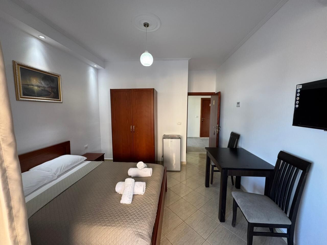 A hotel room with a bed, a desk, a chair, a wardrobe, a television, and a painting on the wall.