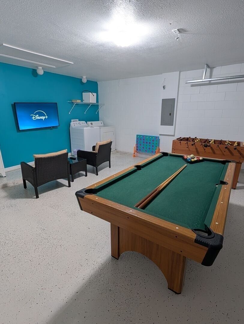 Een pooltafel met omliggende stoelen, een televisietoestel met het Disney+ logo en een wasruimte.