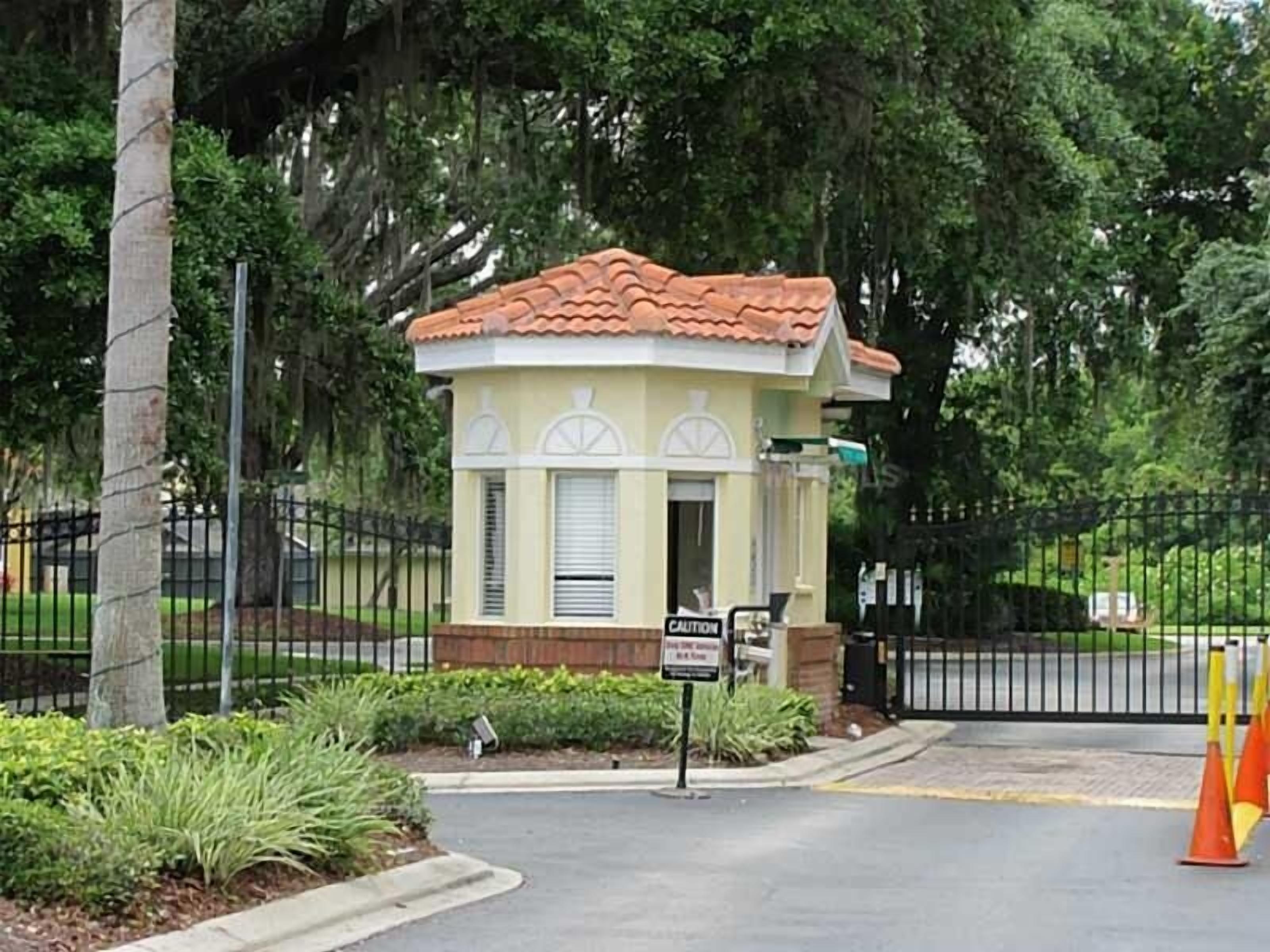 A small, single-story building with a tiled roof and a beige facade, surrounded by a black metal fence and lush greenery.
