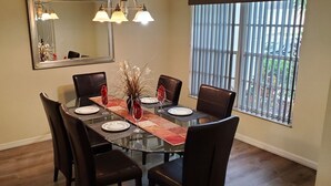 Uma sala de jantar com mesa de vidro, seis cadeiras, um vaso com flores e uma janela com persianas verticais.
