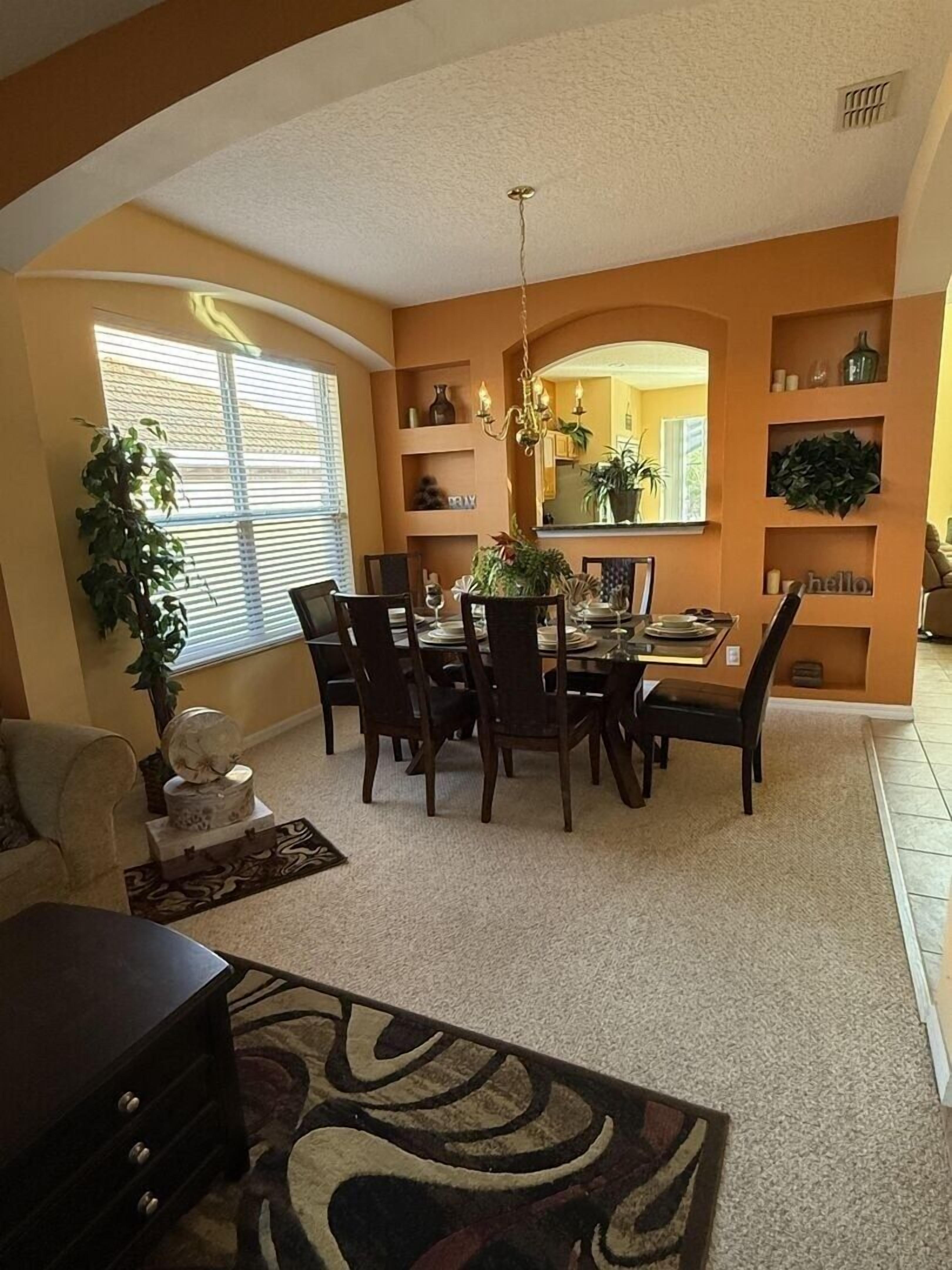 A dining area with a table set for six, surrounded by chairs, a chandelier, and shelves with decorative items.