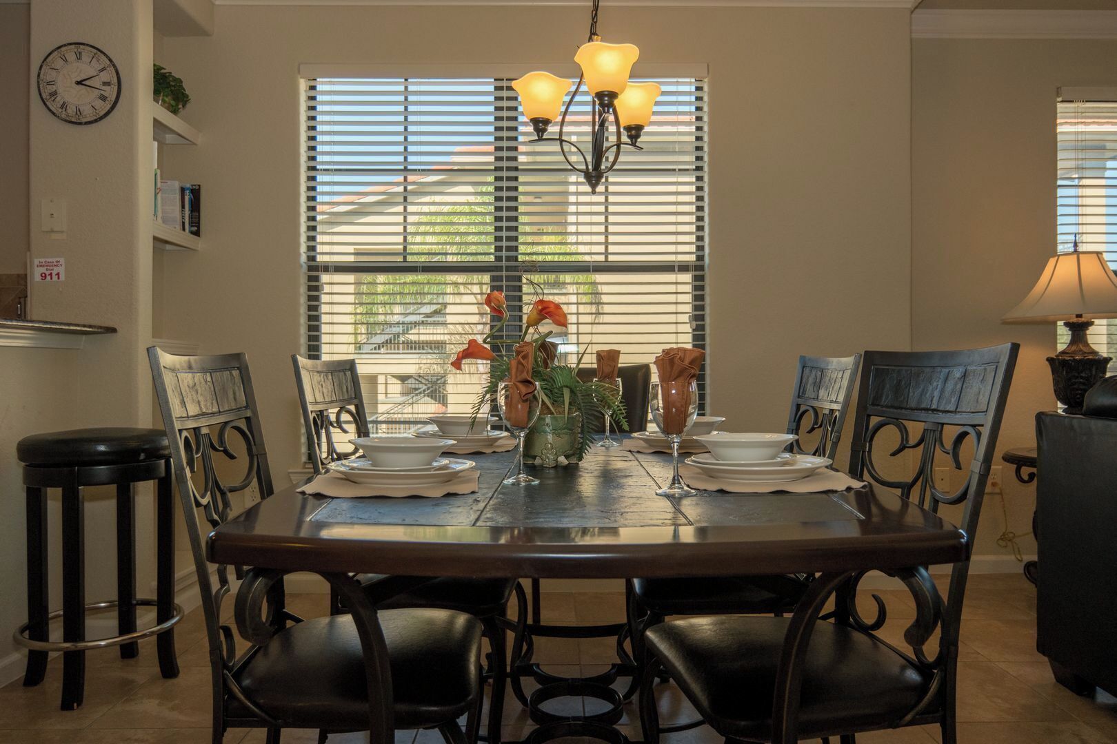 A dining room with a wooden table set for four, surrounded by chairs. A chandelier hangs above, and a window with blinds is visible in the background.