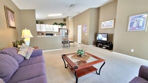 A living room with a purple sofa, a coffee table, and a TV stand with a flat-screen TV. There is a kitchen area in the background with cabinets and a refrigerator. - HL740BD 4 Bedroom Villa in Hampton Lakes 4 Home (Davenport)