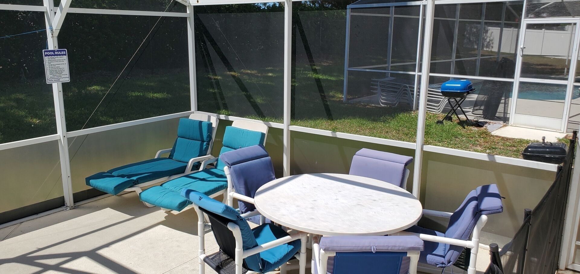 A covered patio area with blue cushioned chairs and a round table, a pool with a sign indicating rules, and a view of a backyard with a grill.