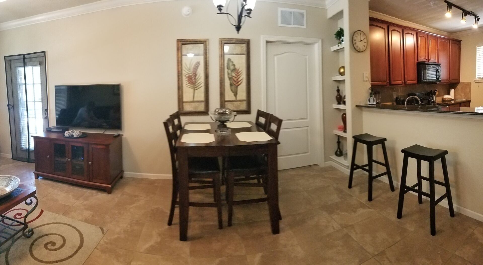 A dining area with a table and chairs, a television, and a kitchen area with cabinets and stools.
