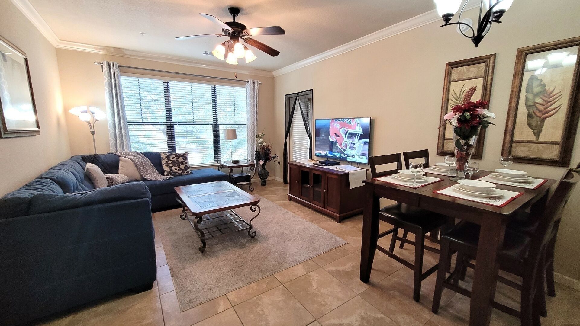 A living room with a dark blue sofa, a coffee table, a TV on a wooden stand, and a dining area with a table set for four.
