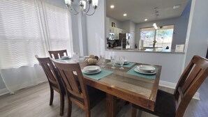 A dining area with a wooden table set for four, surrounded by chairs. A kitchen is visible through an open doorway, and the room has a modern design with light-colored walls and a chandelier. - Sol1520sc 6 Bed Solana Holiday Villa 6 Bedroom Home (Davenport)