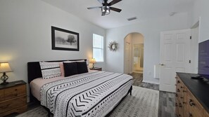 Une chambre à coucher avec un lit, des tables de chevet, un ventilateur de plafond, un tableau encadré au mur et une porte donnant sur une autre pièce.