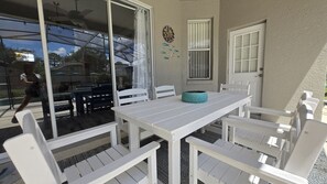 Une terrasse avec une table et des chaises blanches, une horloge décorative et une porte vitrée donnant sur un espace piscine.
