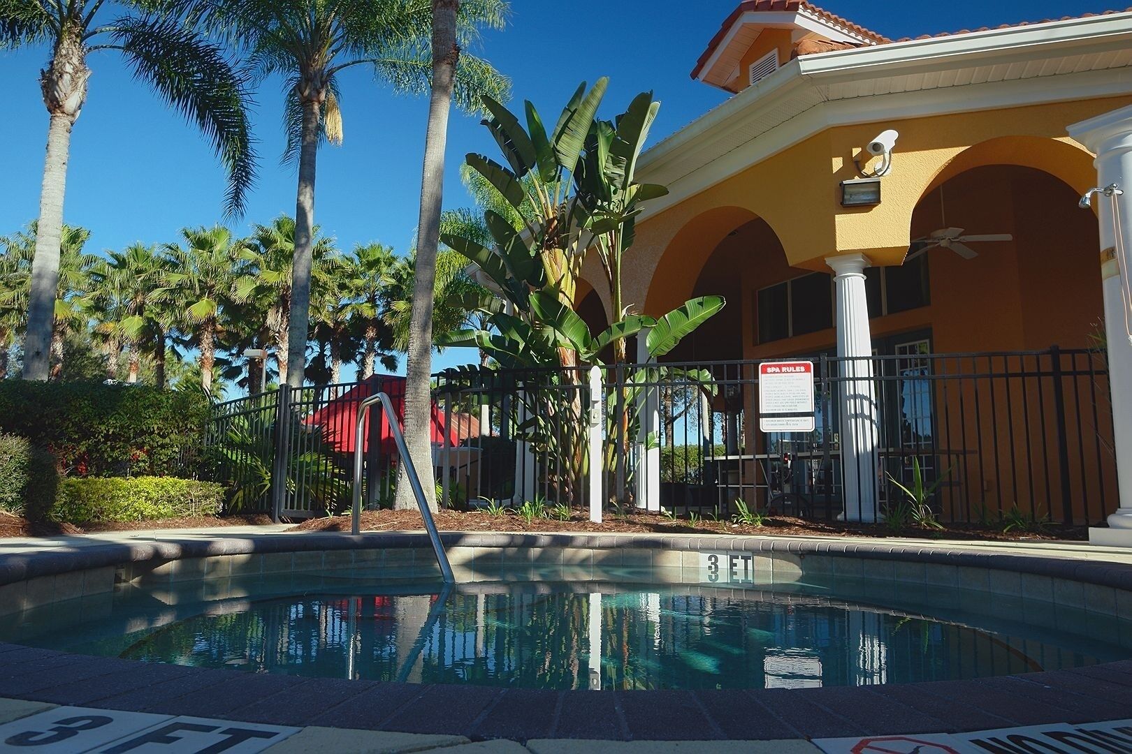A swimming pool with a 3 FT depth sign, surrounded by palm trees and a building with a yellow facade and arched windows.