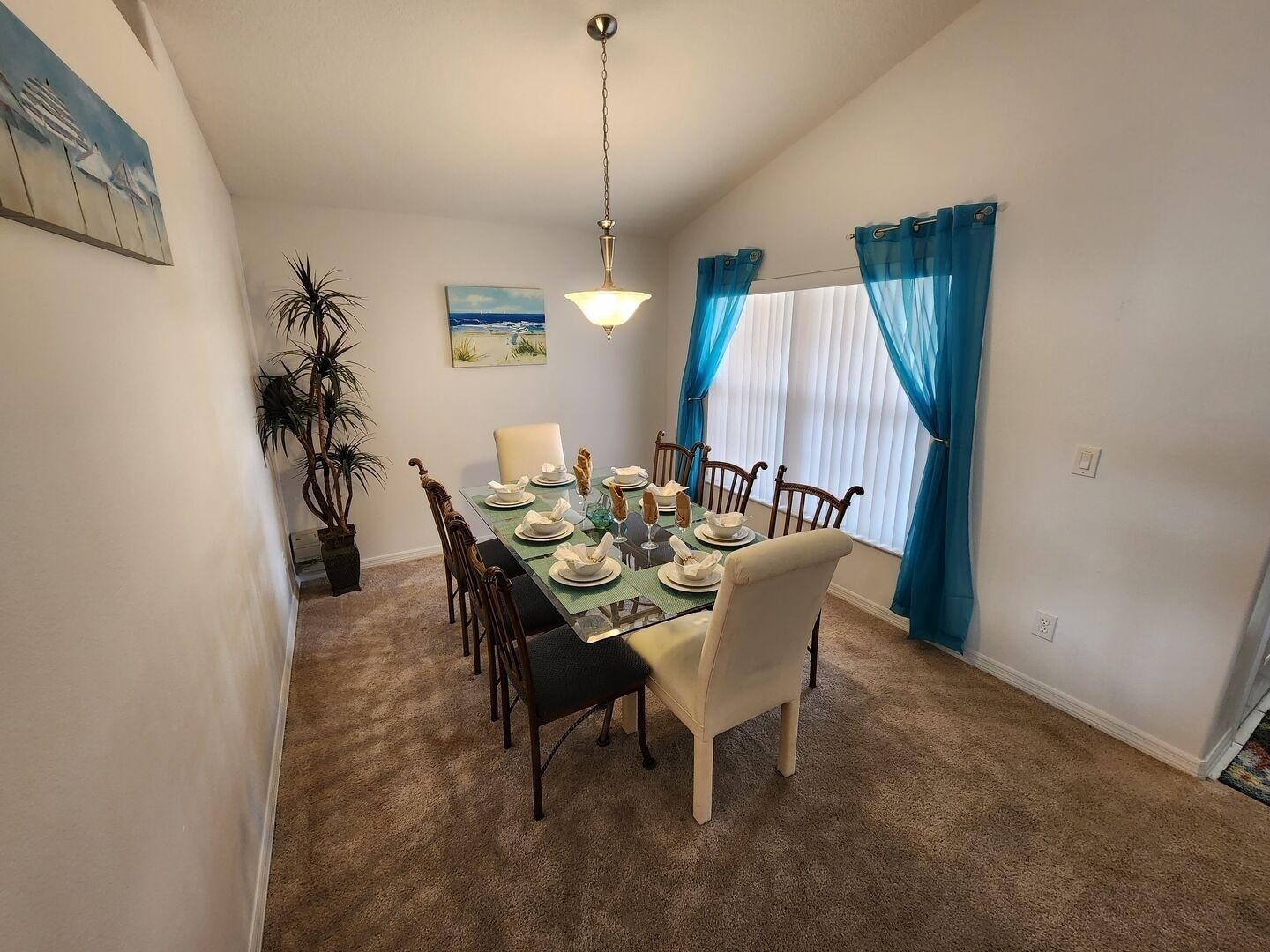 A dining room with a table set for six, surrounded by chairs, a hanging light fixture, and a large window with blue curtains.