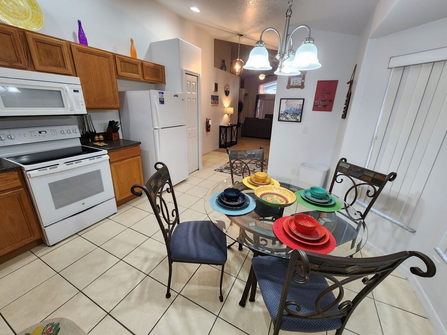 A kitchen with wooden cabinets, a white microwave, a white refrigerator, a stove, and a dining table with colorful plates and blue chairs.