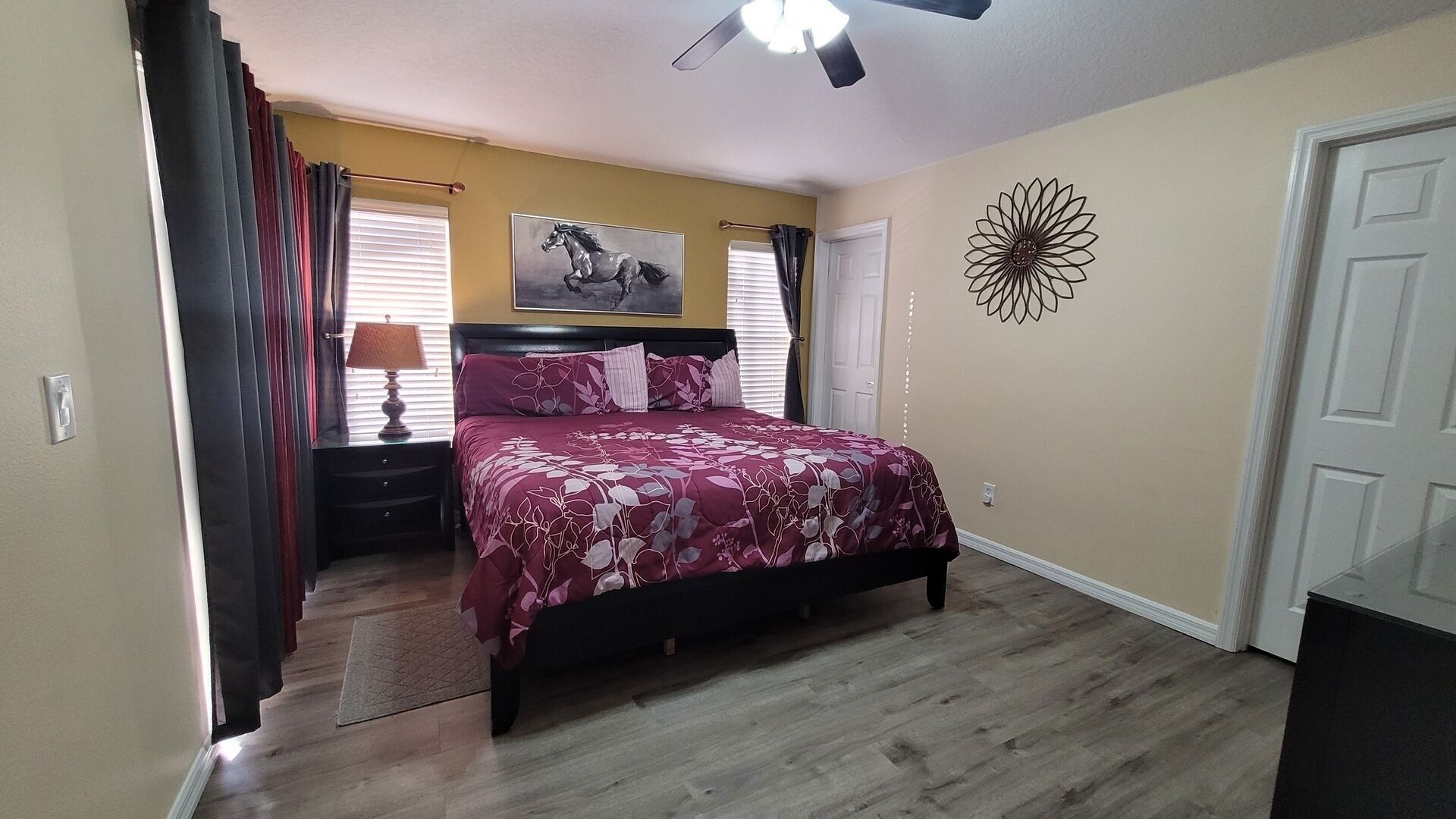 A bedroom with a bed, a nightstand, a lamp, a ceiling fan, a window with curtains, a door, and a wall decoration.