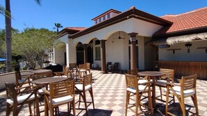 Une terrasse extérieure avec des tables et des chaises en bois, un sol à carreaux et un bâtiment surmonté d’un toit recouvert de tuiles rouges.
