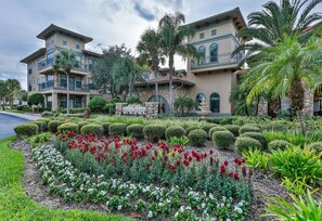 Un hotel de varios pisos con un jardín ajardinado que presenta flores rojas y blancas, palmeras y un letrero de piedra que dice "Bella Piazza".
