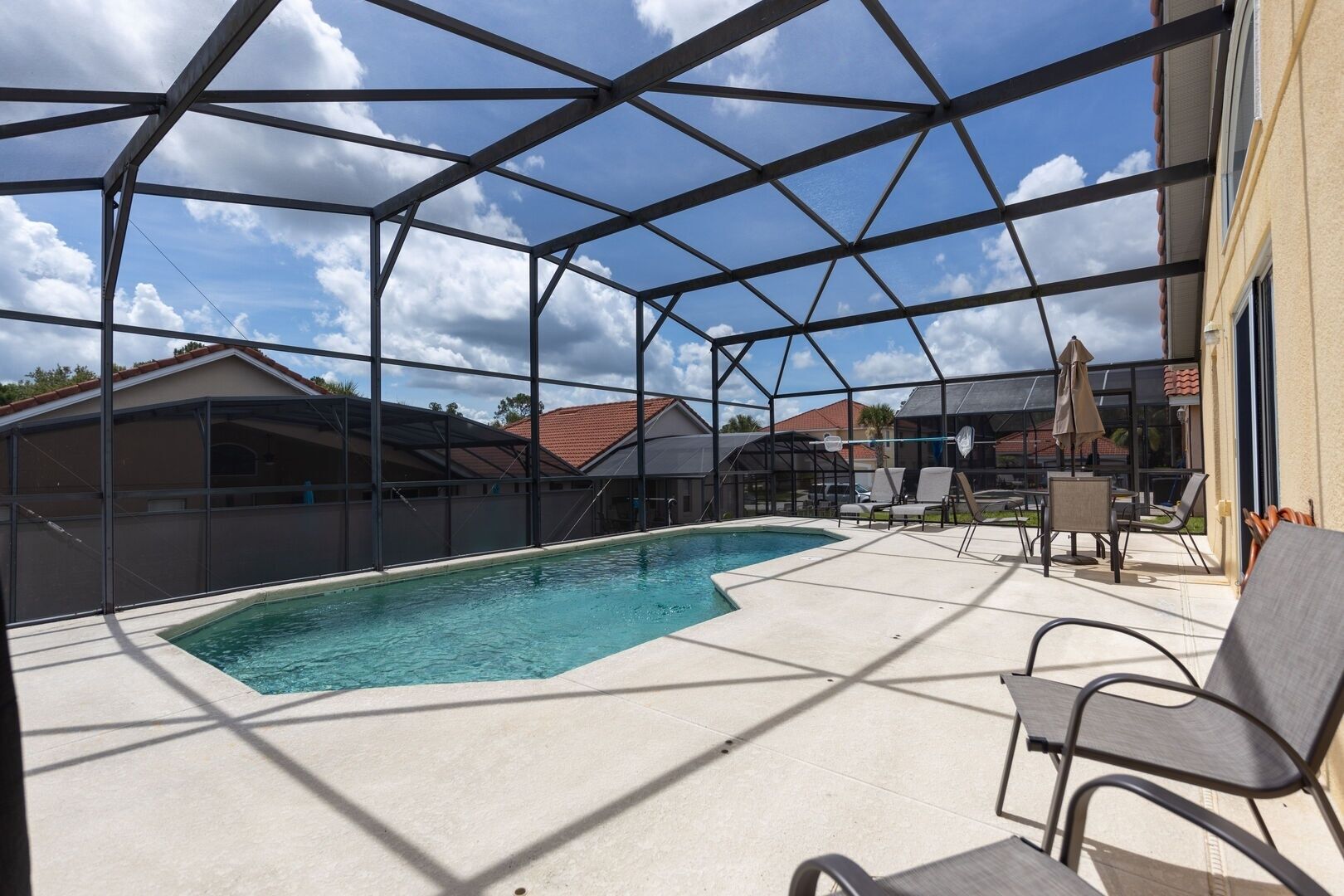 A covered outdoor swimming pool with a clear view of the surrounding houses and a patio area with chairs and umbrellas.