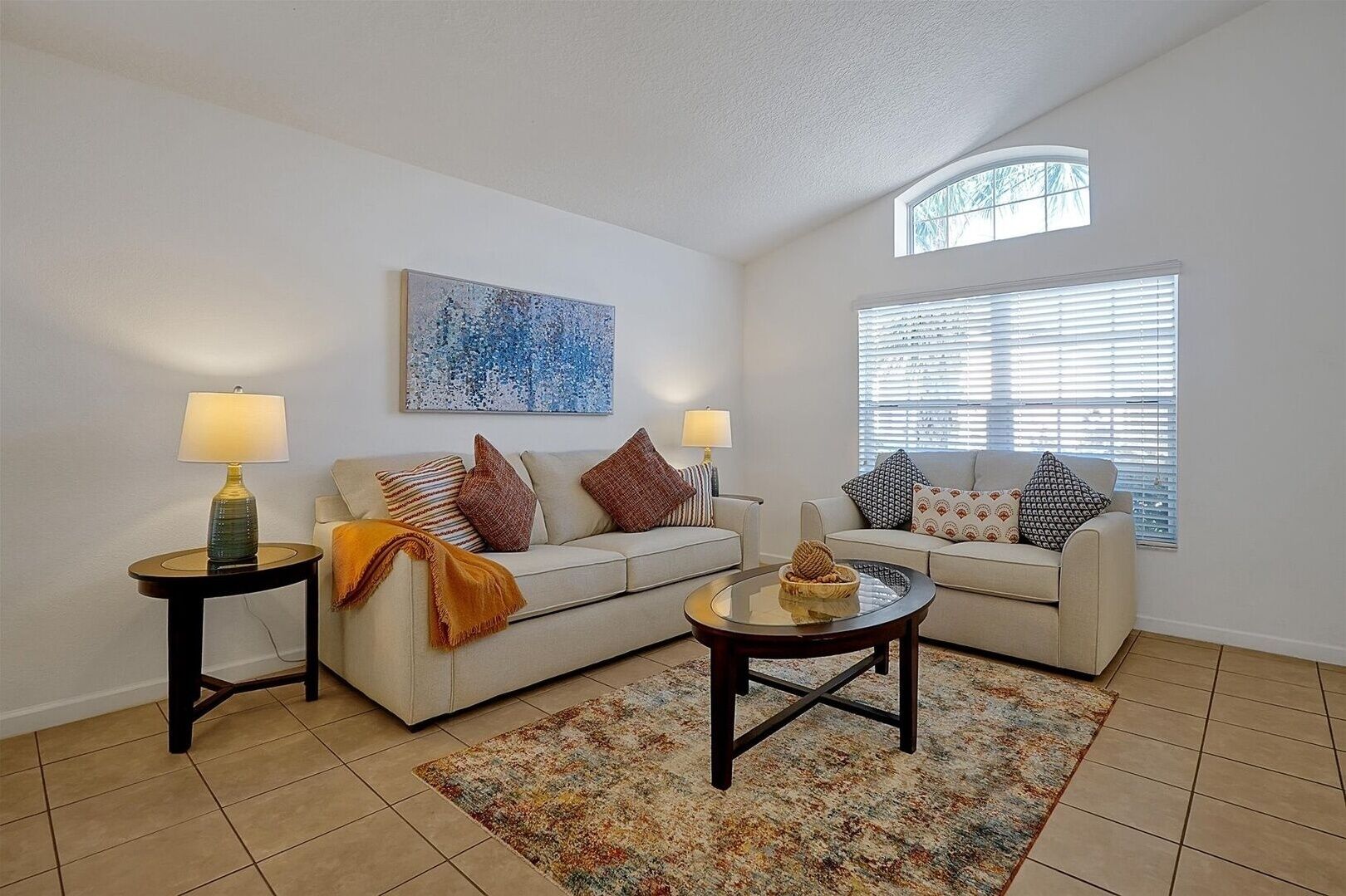 A living room with two sofas, a coffee table, and two lamps. There is a large window with blinds and a ceiling light.