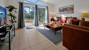 A modern living room with a sofa set, a coffee table, and a TV. There is a large window with curtains, a painting on the wall, and a plant. - Av410vd Relax Retreat Villa In Aviana Resort 4 Bedroom Home (Davenport)
