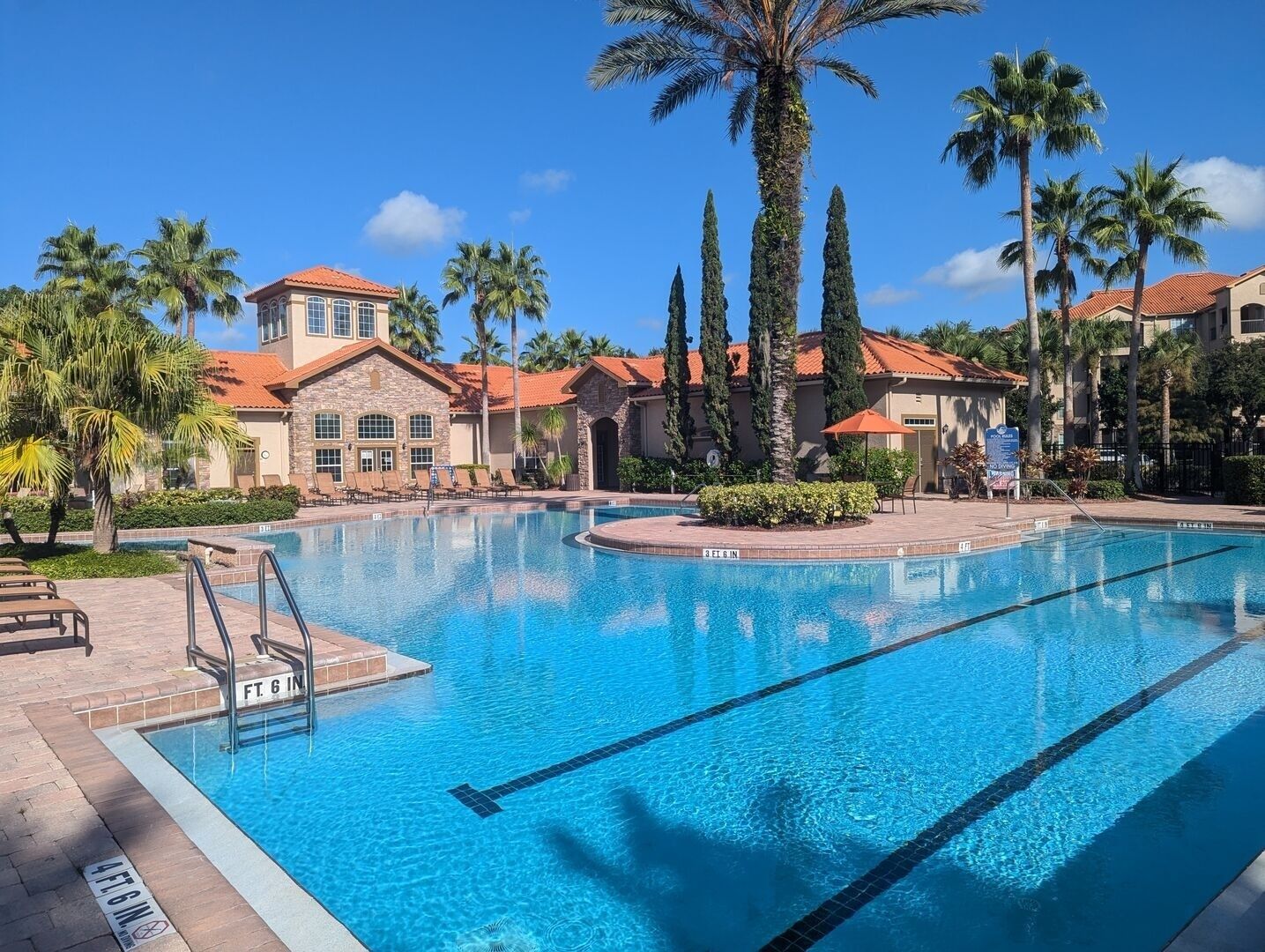 A swimming pool with clear blue water, surrounded by palm trees and a building with a red-tiled roof.