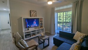 Una sala de estar con televisor, ventilador de techo, sofá, sillón y ventana con vista a follaje.