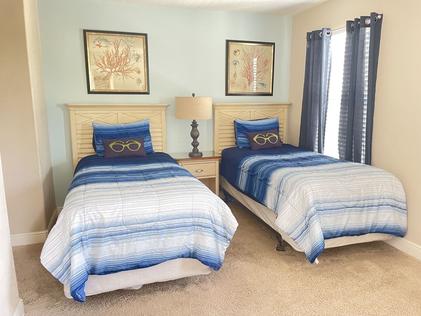 A bedroom with two beds, each with a blue and white striped bedspread, a nightstand with a lamp, and two framed pictures on the wall.
