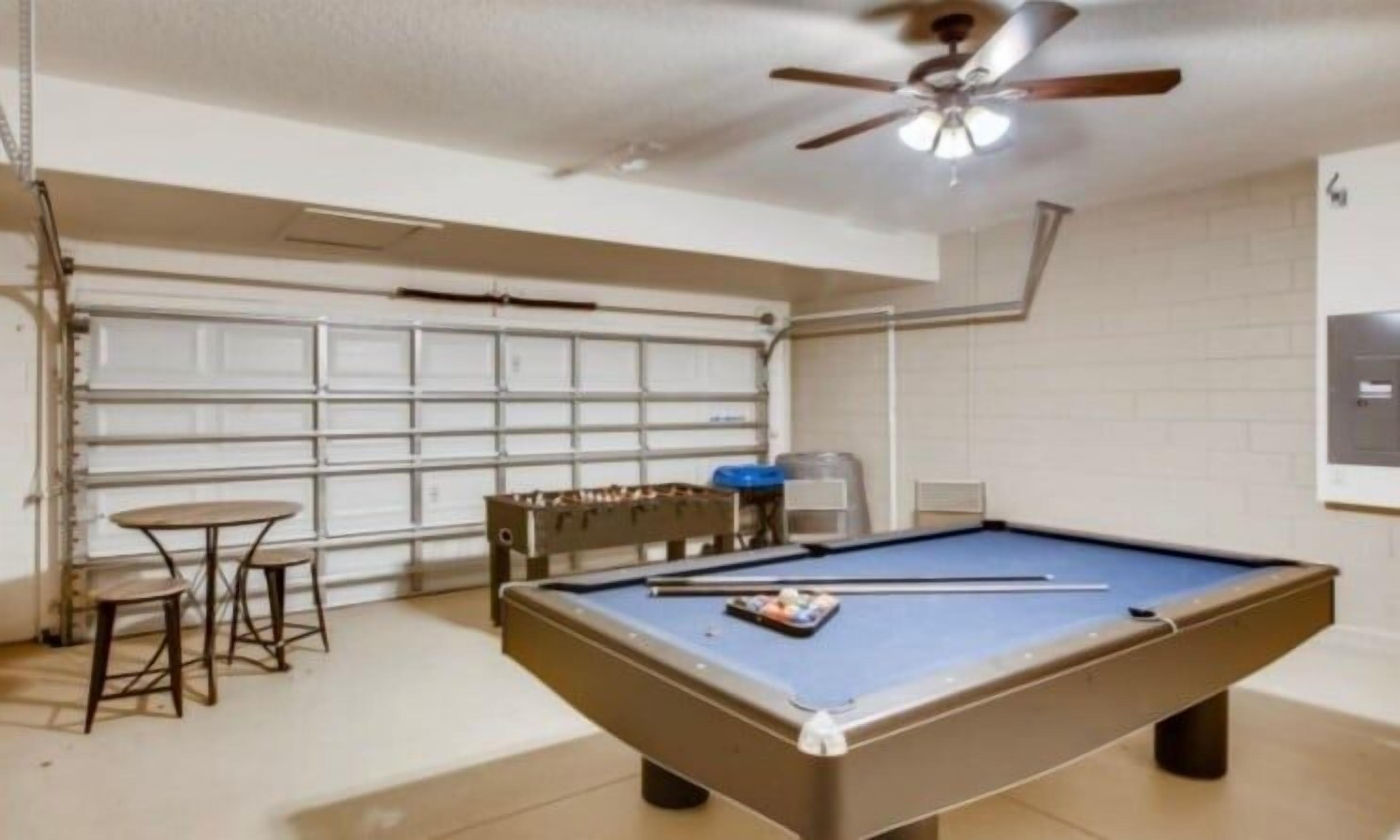 A pool table in a room with a ceiling fan, a small table with chairs, and a garage door in the background.