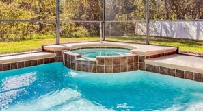Une piscine avec un espace jacuzzi, entourée d’une terrasse couverte et offrant une vue sur un jardin aménagé.