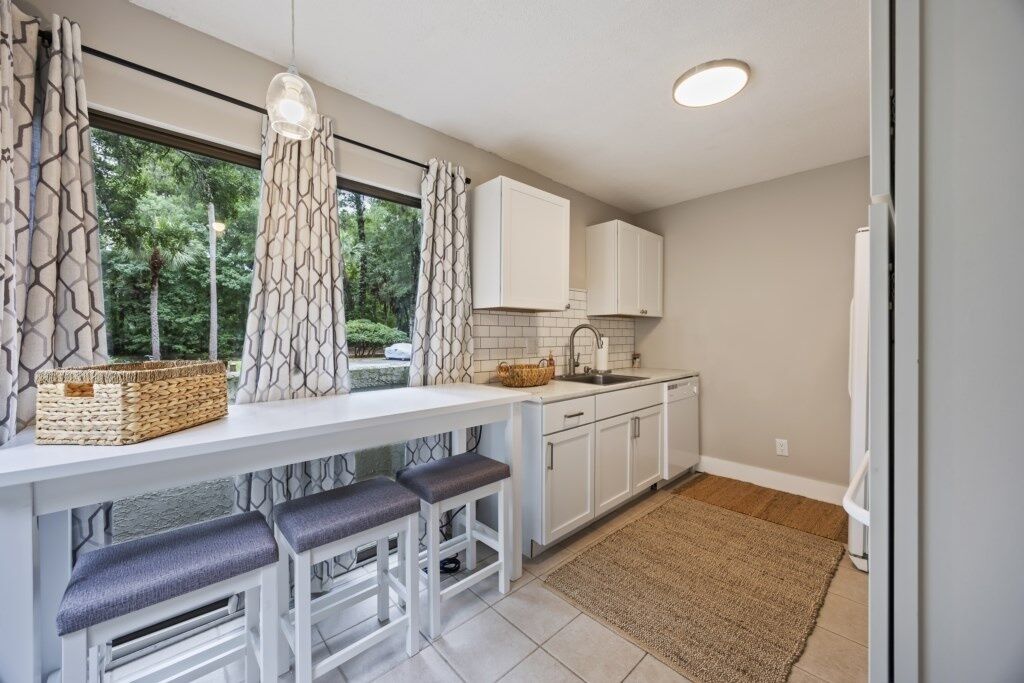 Uma cozinha moderna com armários brancos, um banco embutido com assentos acolchoados, uma janela grande com cortinas estampadas e uma ilha de cozinha com uma cesta e um lustre.
