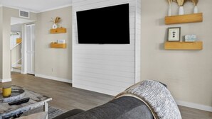 A living room with a mounted TV, wooden shelves with decorative items, a sofa, and a coffee table with remote controls. - Sea Pines - 867 Ketch Court 2 Bedroom Home (Hilton Head Island)