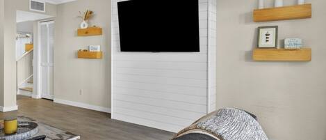Sala de estar con televisor montado, estanterías de madera con objetos decorativos, un sofá y una mesa de centro con controles remotos.