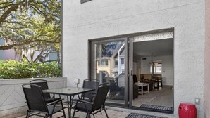 An outdoor patio with a table and chairs, a red trash bin, and a sliding glass door leading to an interior space with a kitchen and dining area. - Sea Pines - 867 Ketch Court 2 Bedroom Home (Hilton Head Island)