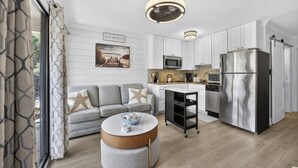 A modern kitchen with white cabinets, a stainless steel refrigerator, a microwave, and a coffee maker. There is a gray sofa with starfish pillows and a round coffee table with a bowl and a candle. - South End - 180 Sea Side Villas 1 Bedroom Condo (Hilton Head Island)