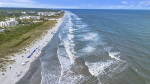 An aerial view of a beach with a row of beach umbrellas and people, a sandy shore, and buildings in the background. - South End - 180 Sea Side Villas 1 Bedroom Condo (Hilton Head Island)