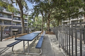 An outdoor picnic area with a long table and benches, surrounded by a metal fence, with trees and a building in the background. - South End - 180 Sea Side Villas 1 Bedroom Condo (Hilton Head Island)