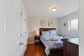 Une chambre avec un plancher en bois, un lit recouvert d’une couvre-lit à motifs, une table de chevet avec une lampe et deux œuvres d’art accrochées au mur.