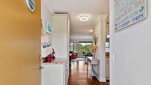 Un couloir étroit avec un plancher en bois, des murs blancs et un luminaire au plafond. Il y a une porte sur la gauche et une fenêtre sur la droite.