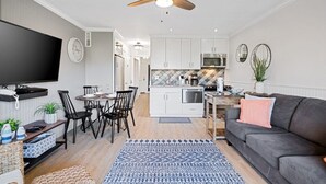 Una sala moderna con un sofá gris, un tapete estampado, un comedor con mesa y sillas, una cocina con armarios blancos y un microondas, y un ventilador de techo.