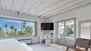 Een slaapkamer met een groot bed, een aan de muur bevestigde televisie, een klein wit kastje en een raam met uitzicht op palmbomen.