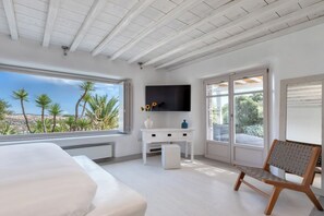 A bedroom with a large bed, a TV mounted on the wall, a small white cabinet, and a window with a view of palm trees. - Lux 9-bdrm professionally managed villa in Mykonos,private pool & sea views (Mikonos)