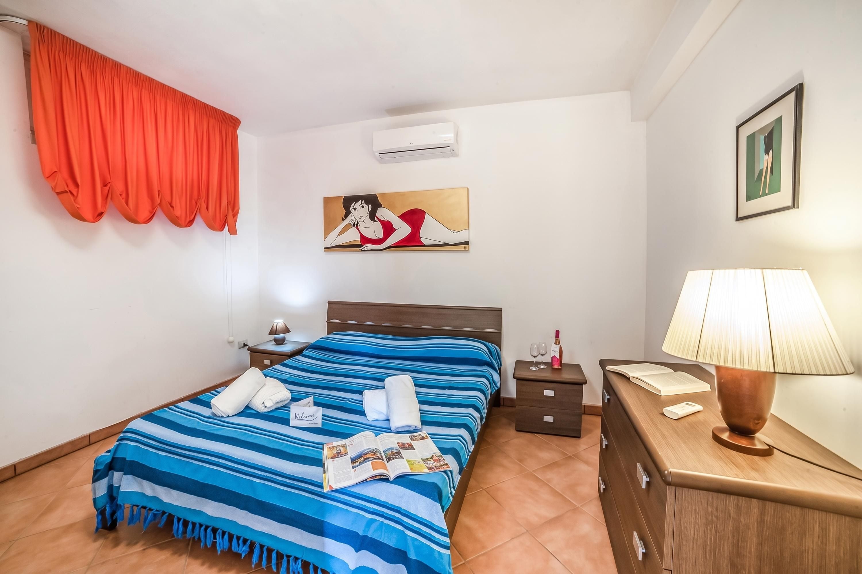 A hotel room with a bed, a nightstand, a lamp, a painting, and a window with curtains.
