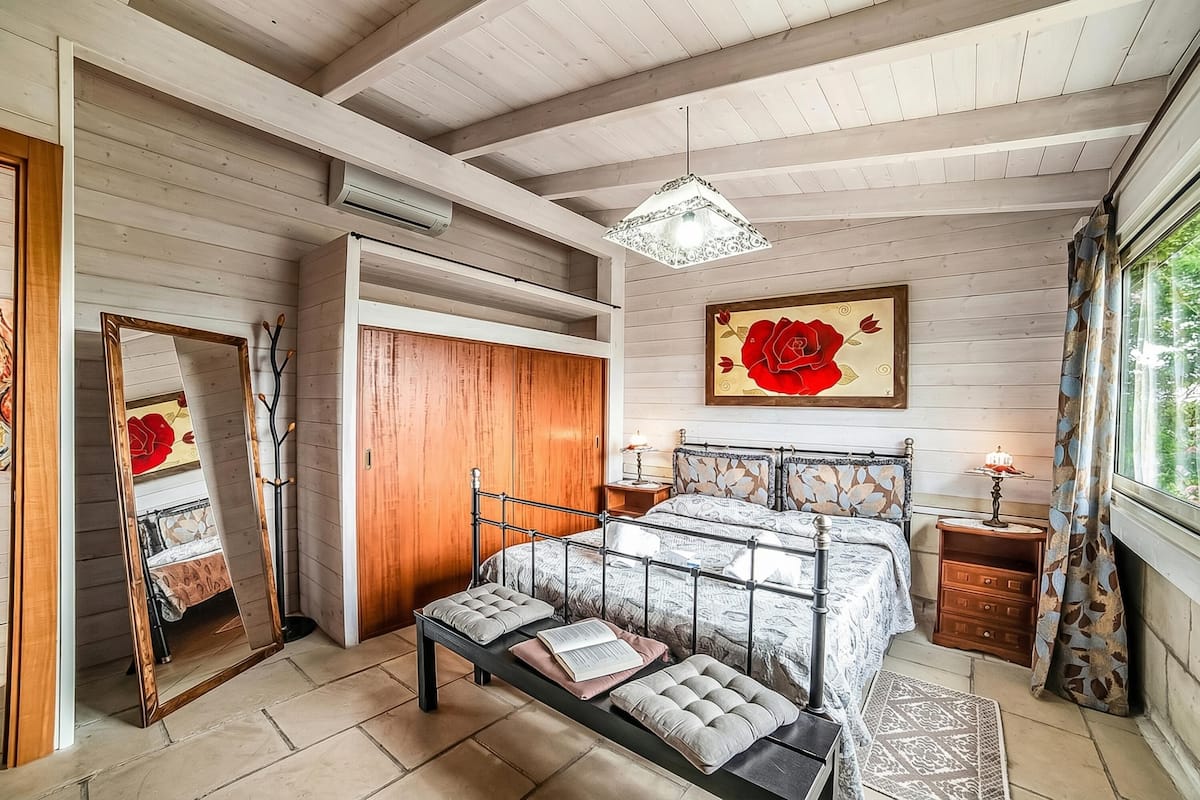 Ein Schlafzimmer mit einem Bett, einer Sitzbank, einem Spiegel, einem Kleiderschrank, einem Nachttisch und einem Fenster mit Vorhängen.