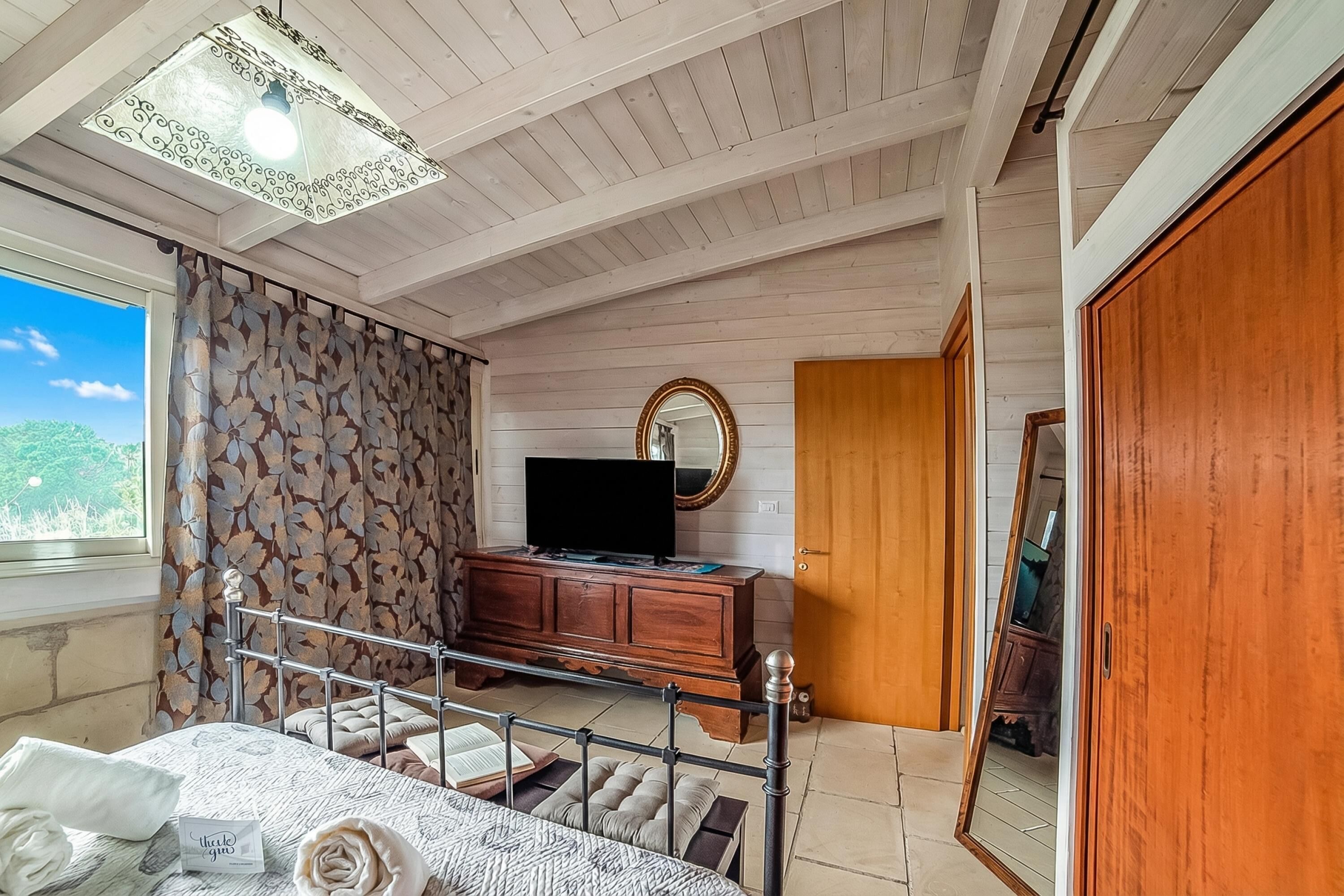 A bedroom with a bed, a dresser, a television, a mirror, and a window with curtains.