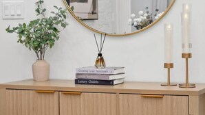 A bedroom with a round mirror, a potted plant, books, candles, and a bed with a brown pillow.