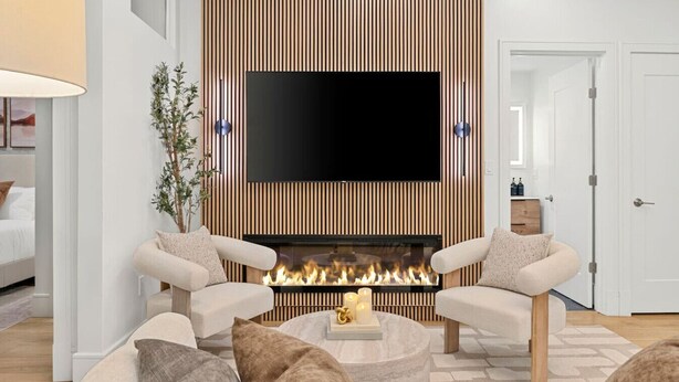 A modern living room with a flat-screen TV mounted on a wooden wall, a fireplace with a visible fire, two armchairs, and a coffee table.